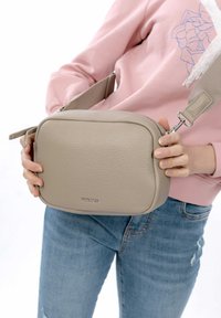 Beige Leder-Crossbody-Tasche mit glatter Textur, rechteckiger Form, silbernem Reißverschluss und Logo auf der Vorderseite. Verstellbarer Riemen sichtbar.