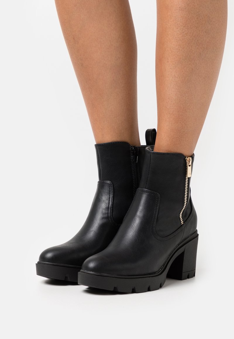 Bottes courtes noires en cuir lisse, dotées d'un talon épais, d'une fermeture éclair latérale avec garniture dorée, et d'une semelle extérieure en caoutchouc texturée.