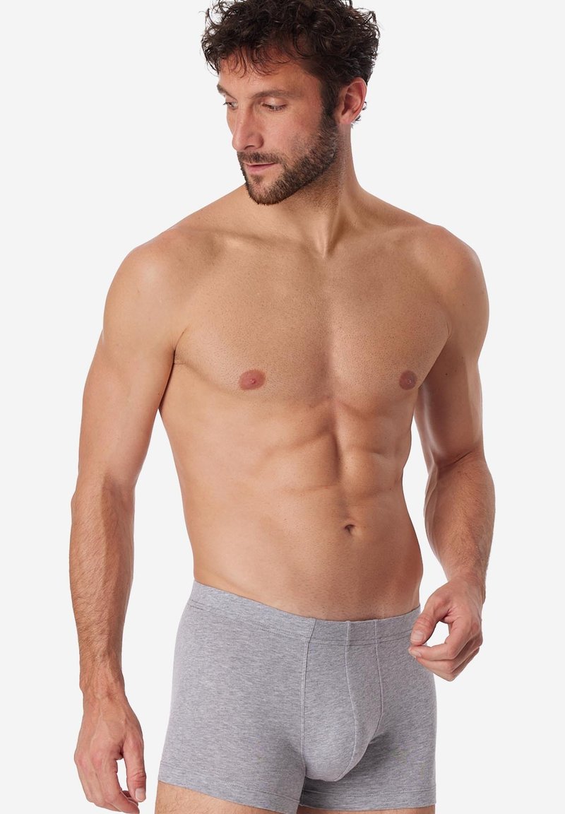 Homme torse nu aux cheveux bouclés portant un boxer gris clair, regardant vers le bas sur sa gauche, sur un fond blanc.