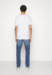 Vit bomull t-shirt med korta ärmar, sedd bakifrån. Bärs med ljusblå jeans och grå sneakers. Enkel design, inga mönster.