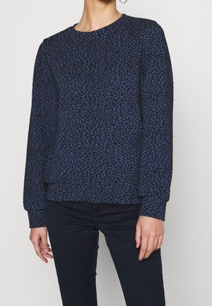 Personne portant un pull à manches longues bleu marine et noir à imprimé léopard, associé à un pantalon foncé, debout devant un fond clair uni.