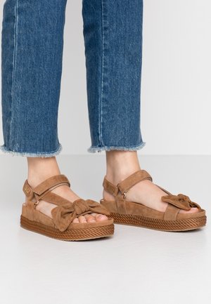 Füße, die braune Wildleder-Plattform-Sandalen mit Knöchelriemen und großen Frontschleifen tragen, kombiniert mit blauen, kurz geschnittenen Jeans mit ausgefranstem Saum.