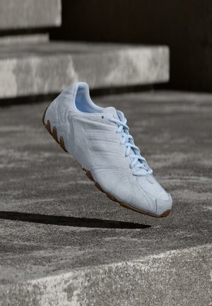 Chaussure de sport bleu clair avec surface texturée et semelle marron, suspendue au-dessus de marches en béton rugueux en extérieur.