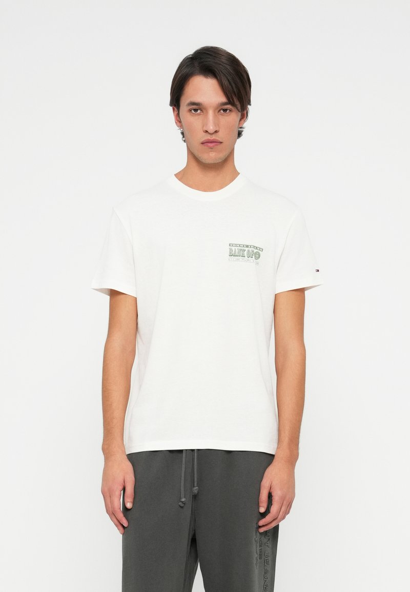 Fiatal férfi fehér grafikás pólóban és sötétszürke zsinóros nadrágban áll egy sima fehér háttér előtt.