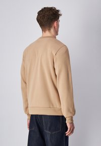Sudadera beige de cuello redondo con puños y dobladillo acanalados. Textura suave, diseño sencillo y un corte relajado. Combinada con jeans de mezclilla azul oscuro.