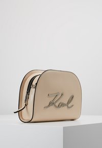 Beige läderväska med rundad form, silverfärgad logotyp med texten "Karl" på framsidan, samt en svart dragkedja som accent vid den övre kanten.