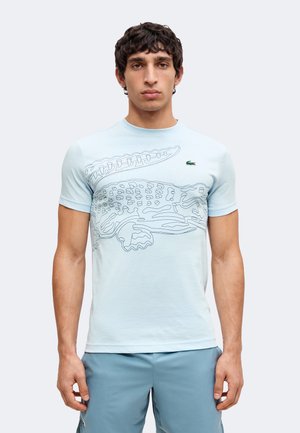 Man wearing light blue t-shirt with large outlined crocodile design and small green logo on the chest, paired with blue shorts.