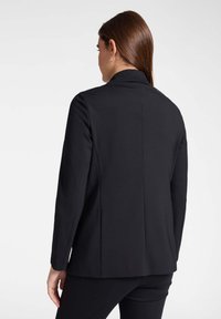 Giacca nera su misura con rever a lanca, maniche lunghe e una texture liscia. Presenta una singola fessura sul retro e una silhouette aderente.