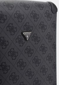 Valigia nera con un motivo testurizzato e logo sottile. Presenta un accentuazione in hardware triangolare nera con marchio "GUESS".