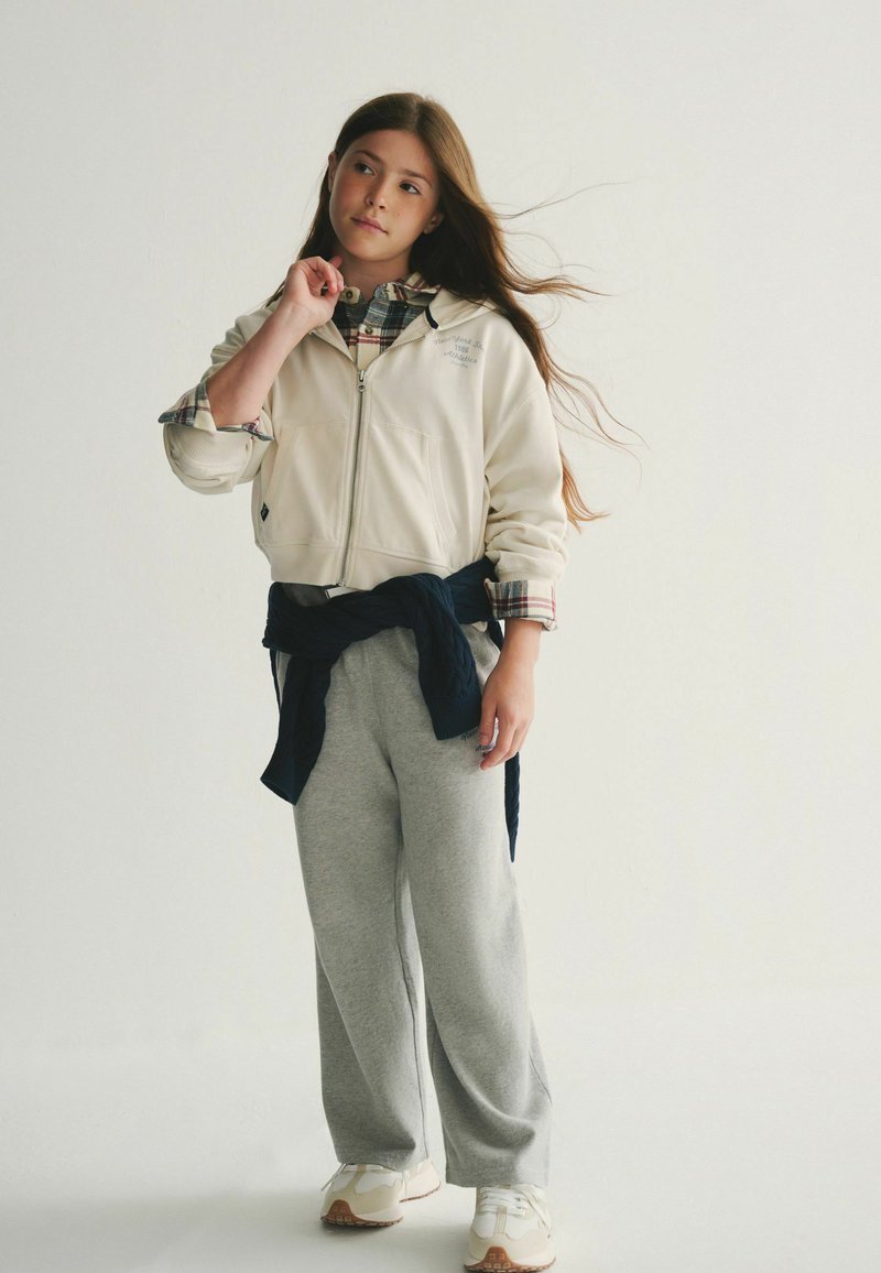 Lichtbeige zip-up jack over een geruite shirt, grijze sweatpants en witte sneakers. Accessoires omvatten een donkerblauwe trui die om de taille is geknoopt.