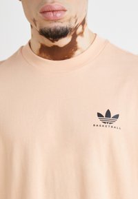 T-shirt de algodão em tom pêssego com gola arredondada e logo preto "BASKETBALL". Textura suave e mangas curtas.