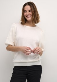 Pull en maille crème à manches courtes avec des manches à volants côtelées, encolure ronde et coupe décontractée, accompagné d'un jean noir.