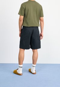 Green cotton t-shirt, black cargo shorts with pockets, white socks, and brown clogs, viewed from the back against a light blue background.