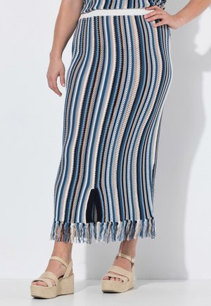 Frau trägt einen langen, gestreiften Strickrock mit Fransen am Saum und beigen Plateau-Sandalen, steht mit einer Hand auf der Hüfte.