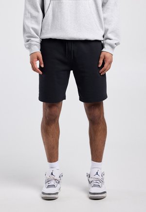 Man draagt zwarte short met trekkoord, witte kousen tot aan de kuit, witte en grijze sneakers, en een lichtgrijze hoodie, staand tegen een effen achtergrond.