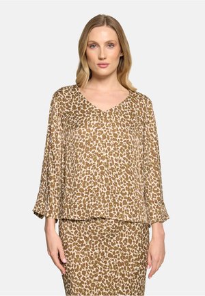 Lyshåret kvinde iført en beige og brun leopardmønstret bluse med lange ærmer og matchende nederdel, stående foran en ensartet lys baggrund.