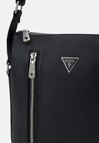 Borsa sintetica nera con finitura testurizzata, dotata di una cerniera argentata e di un logo Guess triangolare. Include una tracolla regolabile per versatilità.