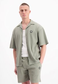 Groene, geweven shirt met korte mouwen en een zwart logo-patch, gecombineerd met bijpassende shorts. Gelaagd over een witte tanktop.