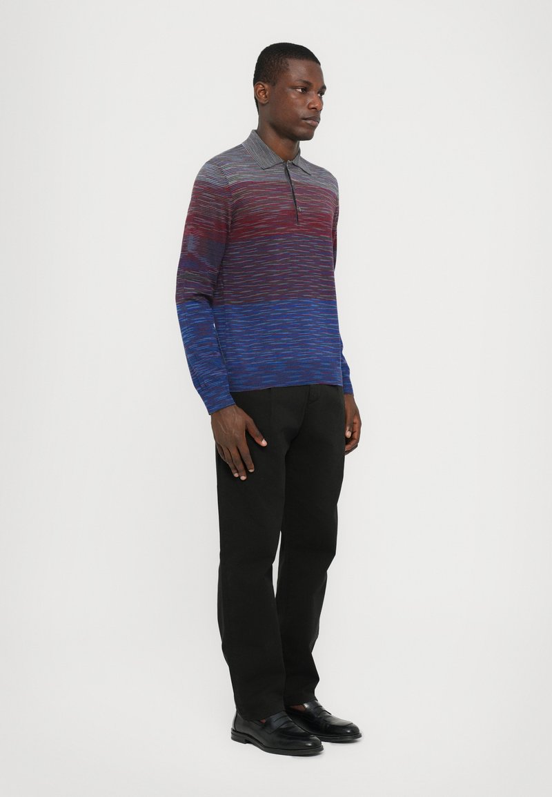 Camisa de manga larga con cuello que presenta un patrón degradado en tonos rojos y azules, hecha de un tejido de punto texturizado, combinada con pantalones y zapatos negros.