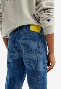 Blue denim jeans featuring a textured graphic pattern, back pockets, and a yellow label peeking from the waistband. Gray sweatshirt partially visible.