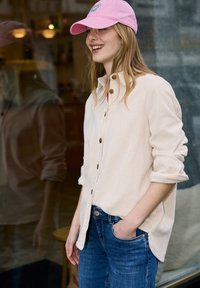 Jeune femme souriante, portant une casquette rose, une chemise crème à boutons avec manches retroussées, et un jean bleu, debout à l'intérieur près d'un mur en verre.