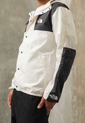 Homme portant une veste The North Face blanche et noire avec logo sur la poitrine et la manche, debout contre un mur texturé neutre.