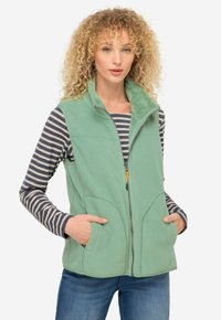 Zelená fleecová vesta se zipem, dvěma předními kapsami a měkkým límcem. Nositá přes pruhovanou dlouhosrstou košili.