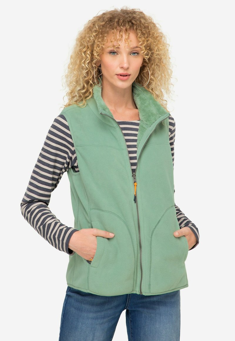 Zelená fleecová vesta se zipem, dvěma předními kapsami a měkkým límcem. Nositá přes pruhovanou dlouhosrstou košili.