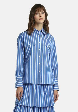 Kobieta ubrana w niebiesko-białą koszulę w pionowe paski z kieszeniami na patki i do tego dopasowaną plisowaną spódnicę na prostym tle.