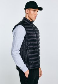 Man in een zwart doorgestikt vest, grijs shirt met lange mouwen, zwarte pet en zwarte broek, staand met handen in de zakken, kijkend naar rechts.