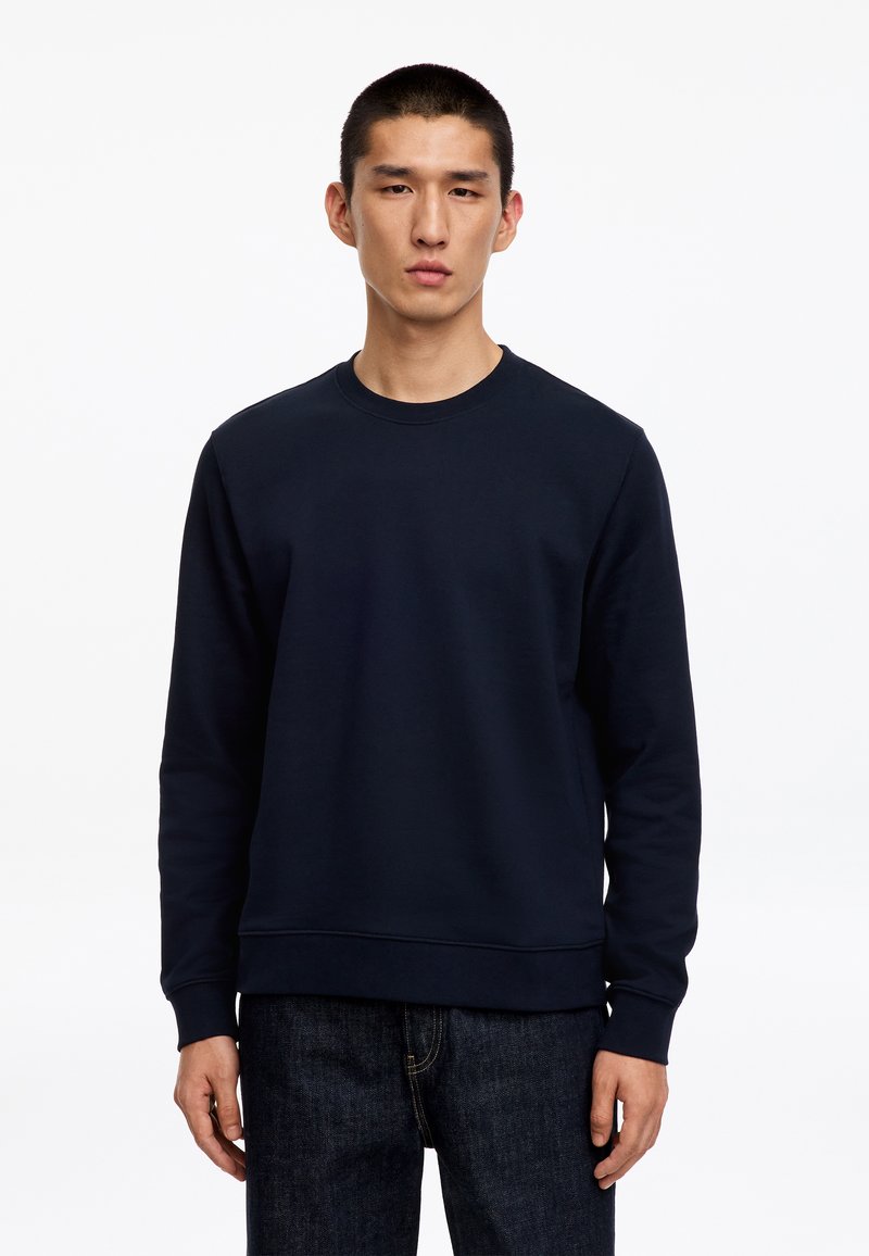 Jeune homme portant un sweat-shirt uni bleu marine foncé et un jean foncé, debout devant un fond blanc.
