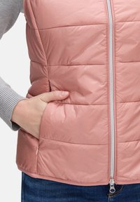 Różowa, ocieplana kamizelka z zamkiem błyskawicznym z przodu, kieszeniami po bokach i poziomym przeszyciem. Tkanina ma gładką, połyskującą fakturę.