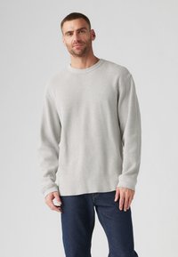 Light gray ribbed long-sleeve shirt with a rounded neckline, paired with dark blue jeans, worn by a smiling man with short hair.