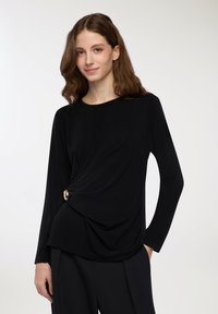 Top nero a maniche lunghe in morbido tessuto con design frontale intrecciato, accentuato da un dettaglio circolare dorato sul lato sinistro.