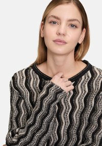 Sweter dzianinowy w kolorze kremowym i czarnym, z falistym wzorem ozdobionym cekinami, okrągłym dekoltem oraz długimi, luźnymi rękawami.