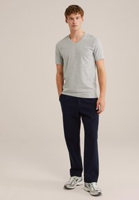 Camiseta gris de cuello en V combinada con pantalones azul marino, el modelo de pie lleva zapatillas deportivas grises y verdes, sobre un fondo claro y neutro.