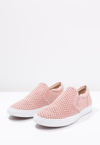 Pár světle růžových slip-on bot s perforovaným svrškem a bílou gumovou podrážkou na jednobarevném bílém pozadí.