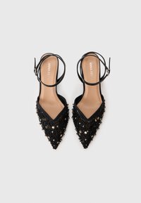 Musta kärkiset tasapohjaiset kengät nilkkaremmillä, koristeltu mustilla paljeteilla ja helmillä, joissa näkyvät ruskeat sisäpohjat ja brändin nimi.