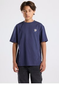 Navy blue short-sleeve t-shirt made of cotton. Features a small logo on the chest. The cut is relaxed with a straight hemline, paired with black pants.