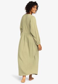 Olijfgroene, lange mouw maxi-jurk van textuurstof met een getailleerde taille en een vloeiende rok. De achterkant toont een afgeronde halslijn en geplooide details.