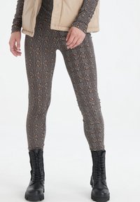 Mustat ja ruskeat kuviolliset legginsit, joissa on geometrinen malli, yhdistettyinä vaaleanbeigeen toppa-aliivihousuun ja mustiin nauhalla varustettuihin maihinnousukenkkiin.