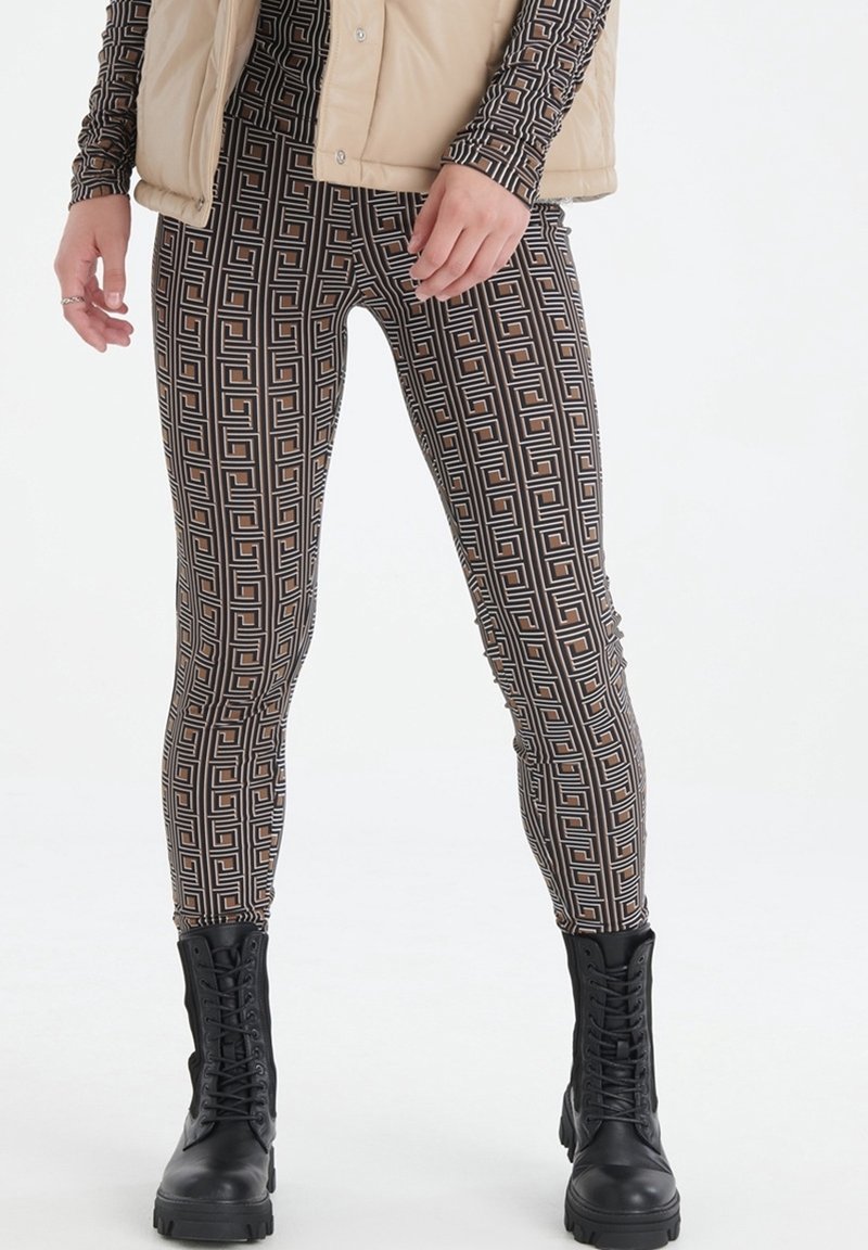 Mustat ja ruskeat kuviolliset legginsit, joissa on geometrinen malli, yhdistettyinä vaaleanbeigeen toppa-aliivihousuun ja mustiin nauhalla varustettuihin maihinnousukenkkiin.