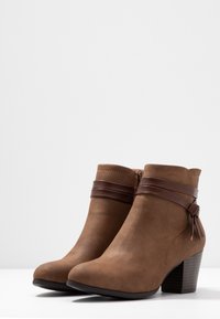 Brązowe zamszowe botki za kostkę z wąskim obcasem, ozdobione brązowym skórzanym paskiem i akcentem w postaci kokardy. Gładkie wykończenie z przeszyciami.