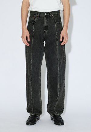 Pantalones vaqueros de mezclilla negra con talle alto, pierna ancha y costuras en contraste. Bolsillos delanteros con herrajes visibles, combinados con zapatos negros.