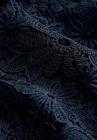 Tmavě modrá látka s propracovanou květinovou výšivkou z texturovaných nití. Vzory zahrnují vířivé motivy a jemné, detailní šití.