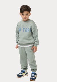 Sudadera verde claro con "BROOKLYN" y "NEW YORK" en impresión azul; pantalones de chándal a juego; zapatillas de tendencia en azul, marino y acentos amarillos.