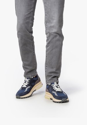Des jambes portant un jean gris et des baskets épaisses bleu marine, gris et beige avec des lacets blancs, se tenant sur une surface blanche.