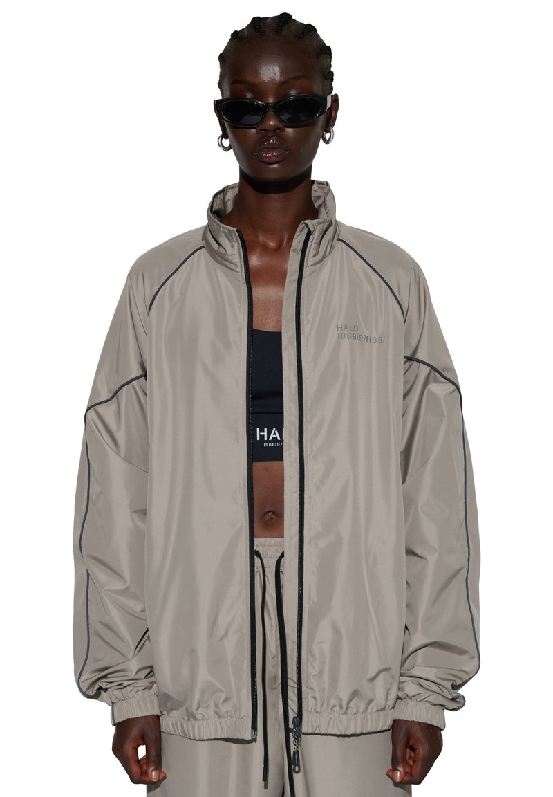 Mørkhudet person med flettet hår bærer en oversize beige jakke, sort sports-bh, matchende bukser, øreringe i ring og sorte solbriller.