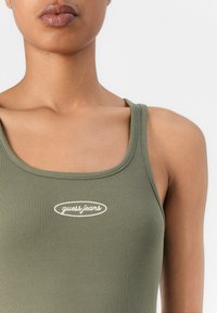 Olīvzaļš ribota žakete ar noapaļotu kakla izgriezumu, uz kuras priekšpuses atrodas balts "guess.jeans" logotips ovālā dizainā.