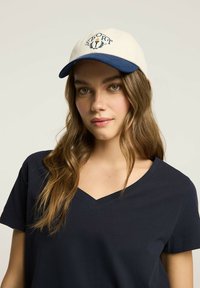 Biała czapka baseballowa z niebieskim daszkiem, z haftowanym napisem "SPORT" i akcentami laurowymi; noszona z ciemnym t-shirtem z dekoltem w kształcie V.